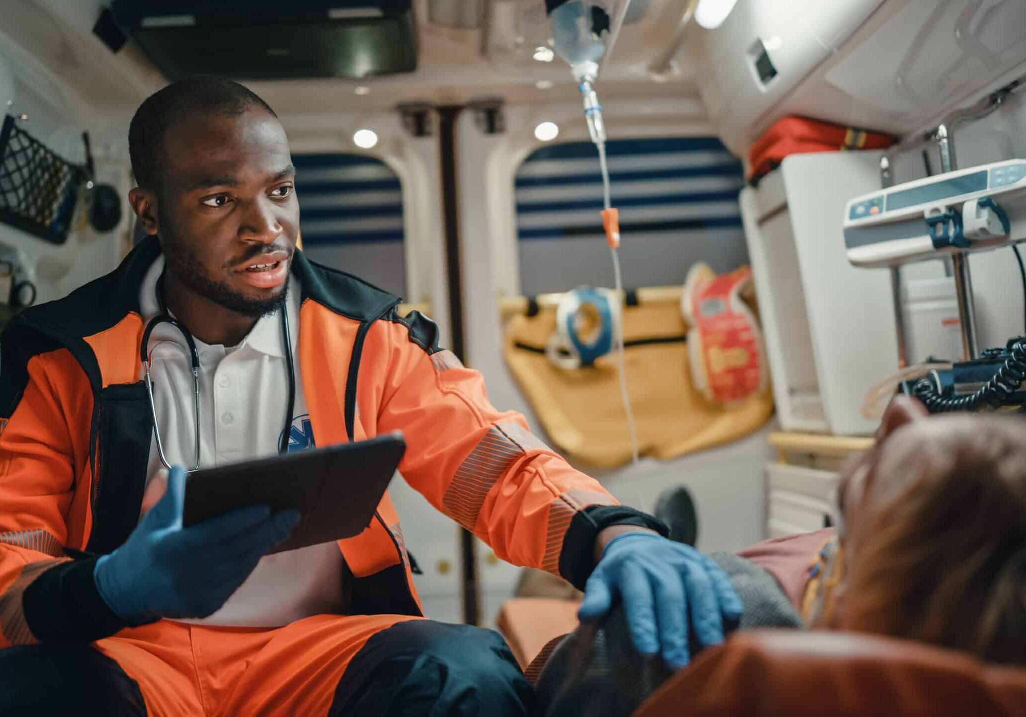 Black African American EMS Professional Paramedic Using Tablet Computer to Fill a Questionnaire for the Injured Patient on the Way to Hospital. Emergency Medical Care Assistant Works in an Ambulance. Black African American EMS Professional Paramedic Using Tablet Computer to Fill a Questionnaire for the Injured Patient on the Way to Hospital. Emergency Medical Care Assistant Works in an Ambulance.